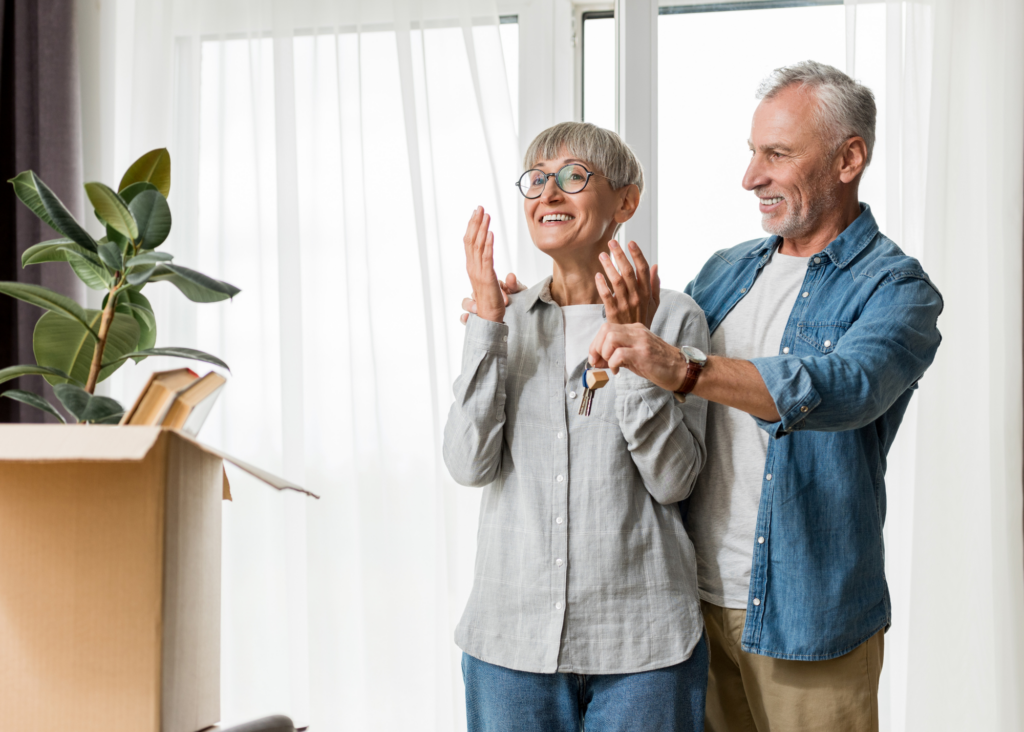 A smiling elderly woman stands near a cardboard box with a houseplant, while a man behind her holds her hands. They appear happy, standing in a bright room with large windows and white curtains, likely enjoying the ease provided by moving services.
