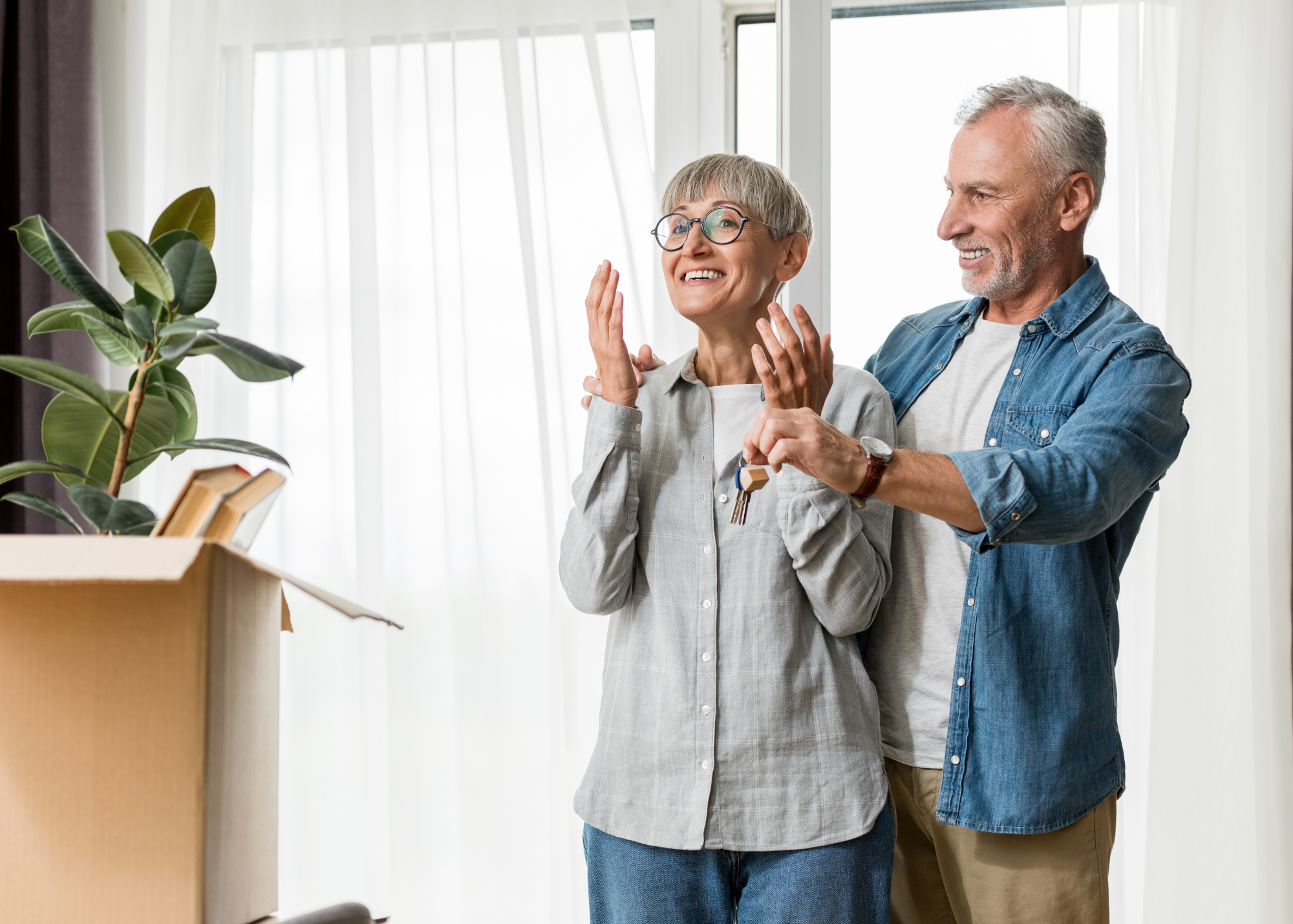 A smiling elderly woman stands near a cardboard box with a houseplant, while a man behind her holds her hands. They appear happy, standing in a bright room with large windows and white curtains, likely enjoying the ease provided by moving services.