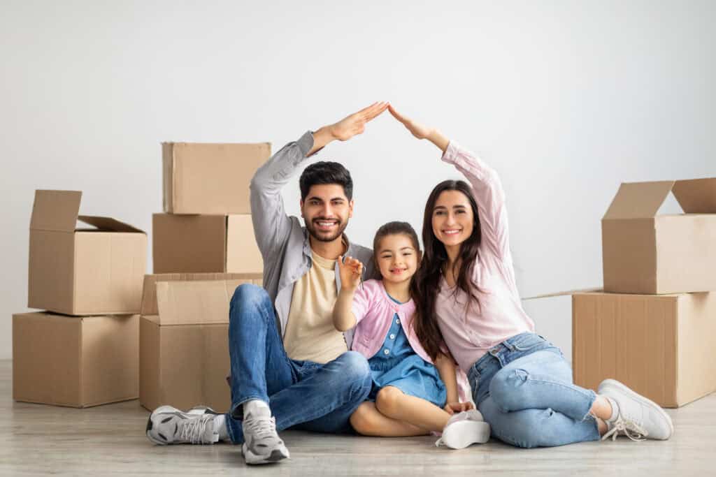 Family of three sitting on the floor, surrounded by moving boxes. Parents are forming a roof shape with their arms over their child.