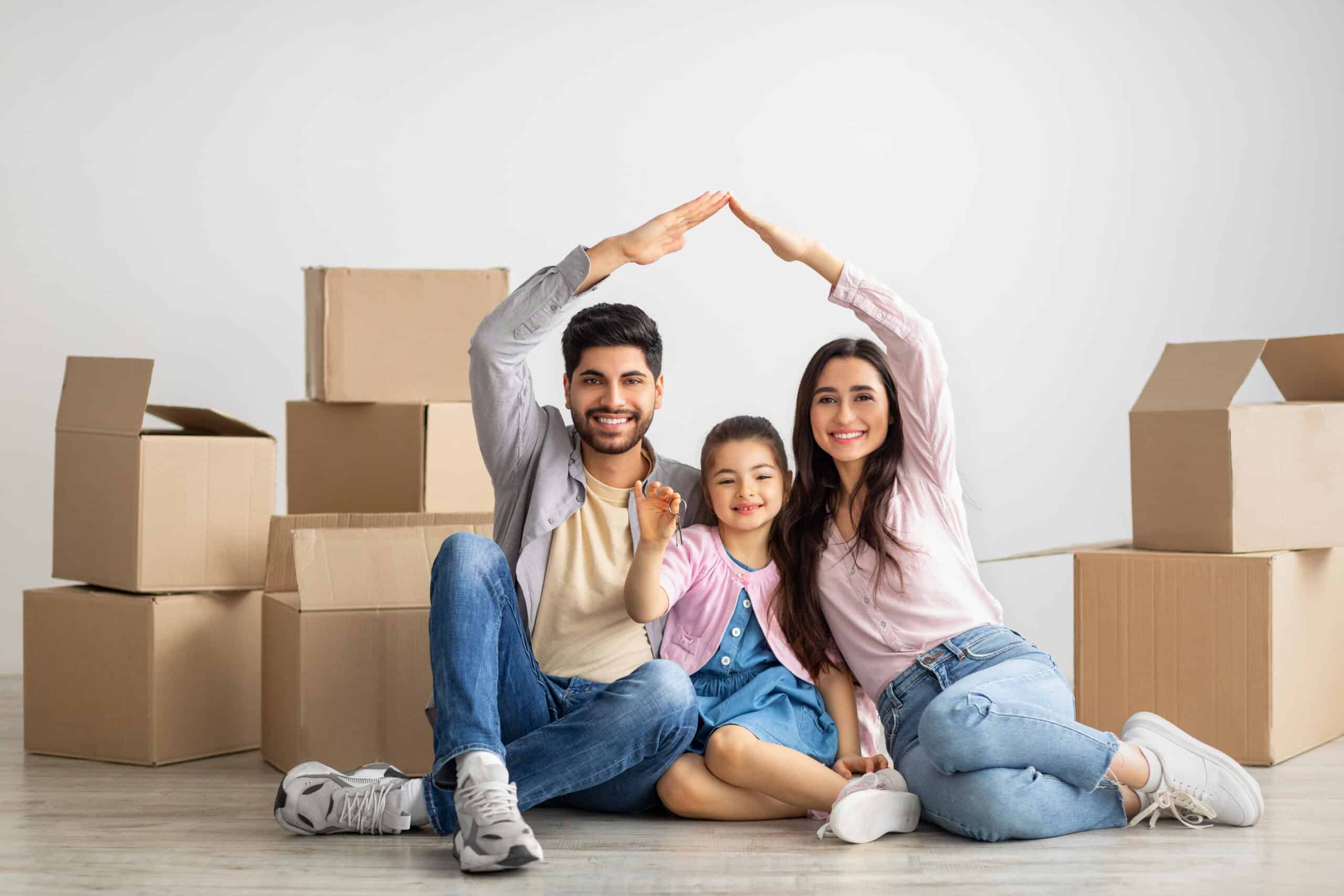 Family of three sitting on the floor, surrounded by moving boxes. Parents are forming a roof shape with their arms over their child.