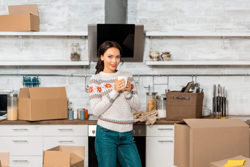 A person in a kitchen holding a mug, surrounded by moving boxes.