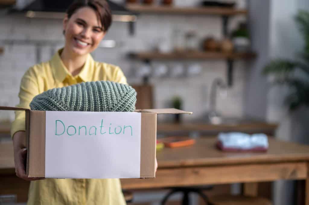 A person holds a donation box containing a knitted item in a kitchen.