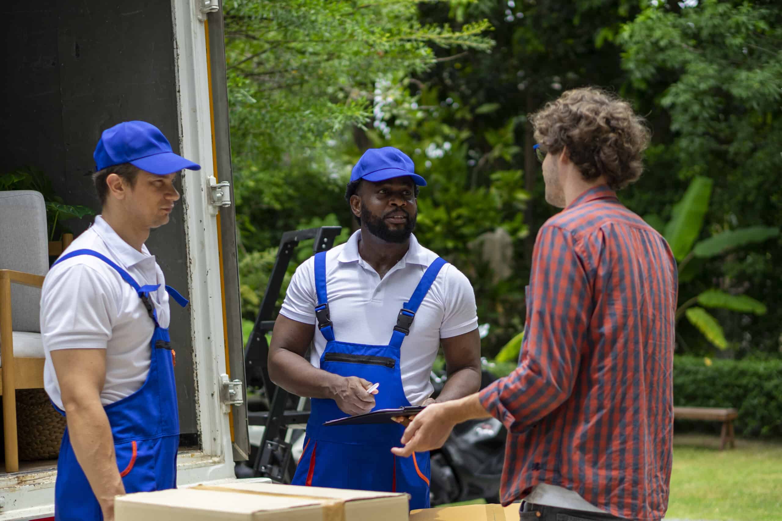 Two delivery workers in blue uniforms stand by a truck talking to a person in a plaid shirt while holding a clipboard.