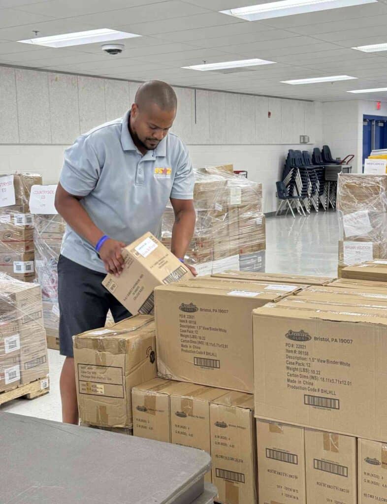 A person in a light gray shirt and shorts stacks cardboard boxes in a room with stacked chairs and more boxes in the background.