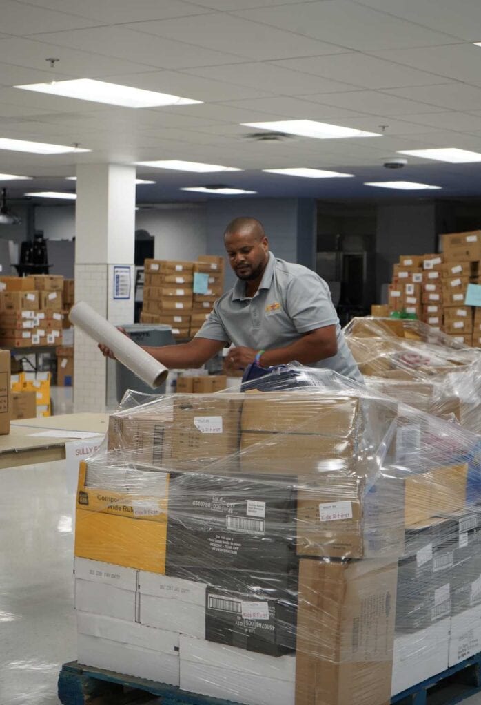 A man in a gray shirt is organizing boxes on a pallet in a warehouse filled with stacked boxes.