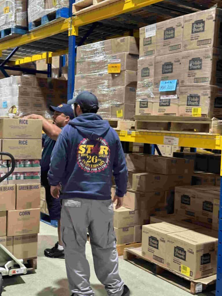 Two workers organize and inspect stacked boxes in a warehouse. One man wears a dark hoodie and cap, while the other wears a blue shirt. Boxes are labeled with items like rice and corn.