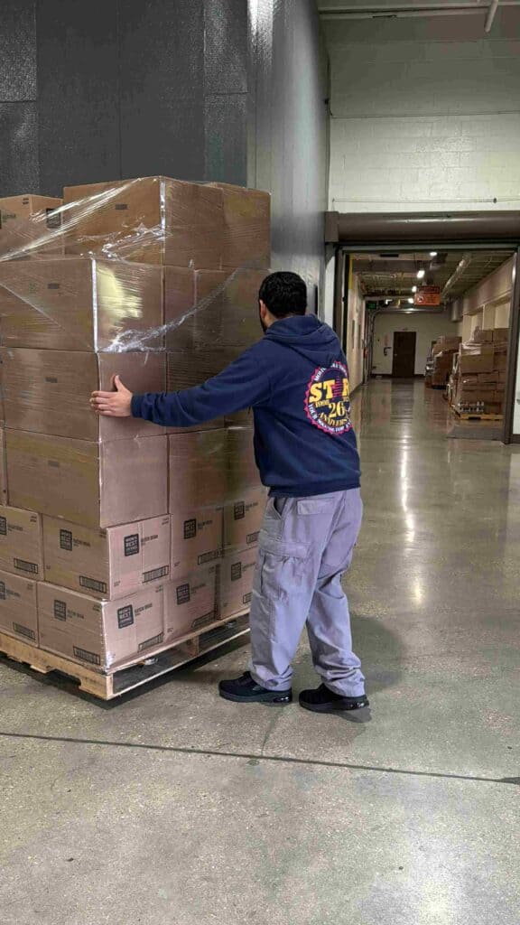 Eine Person, die einen Kapuzenpulli und eine Cargohose trägt, bewegt eine mit Kisten beladene Palette in einer Lagerhalle.