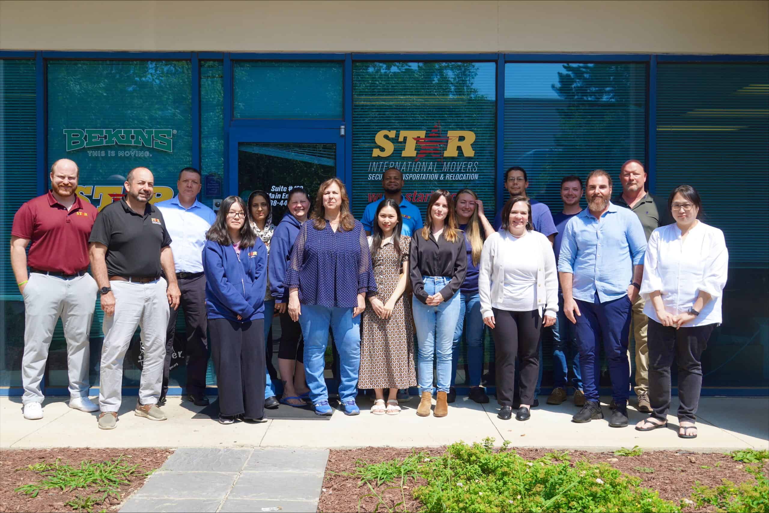 A group of fifteen people stands in a row outside a building with blue windows and signs reading “STAR,” “Starint,” and “BHMS.” They are smiling and facing the camera.